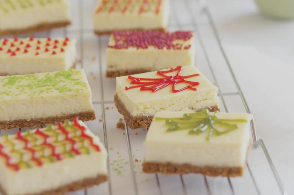 Rectangular cheesecake bars on a cooling rack, each topped with colorful icing designs and sprinkles, creating a festive and inviting appearance.