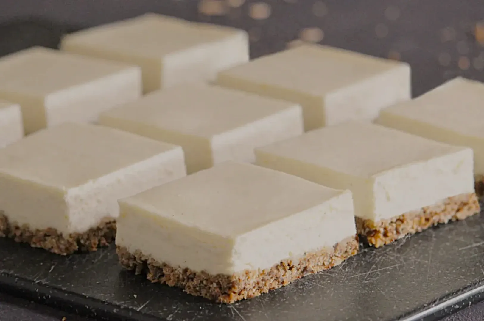 Nine neatly arranged square cheesecake bars sit on a dark slate surface. Each bar features a thick, creamy layer atop a crumbly brown crust.