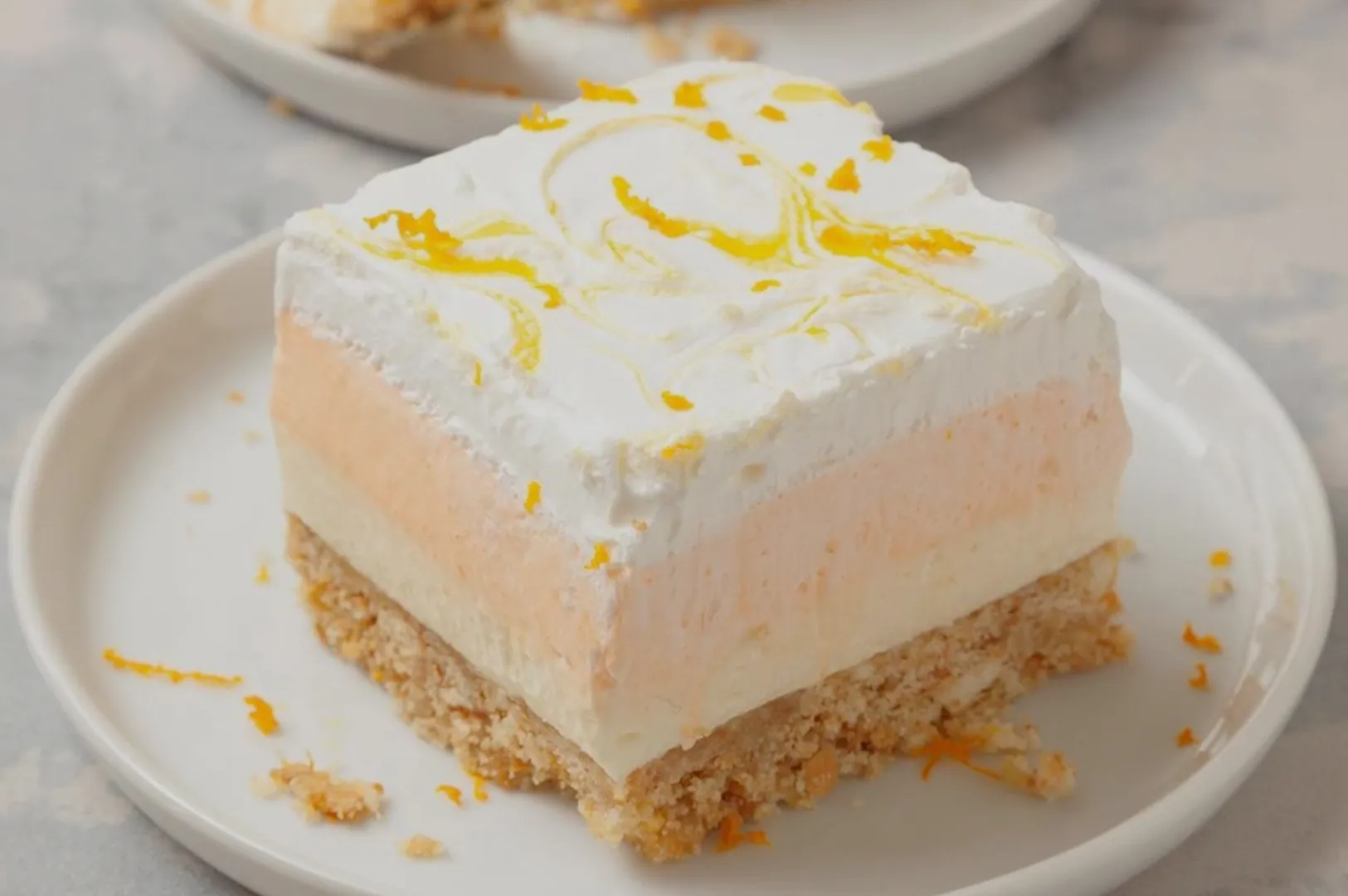 A creamy, layered dessert sits on a white plate. It features a crumbly base, a pale peach and white middle, topped with whipped cream and zesty orange swirls.