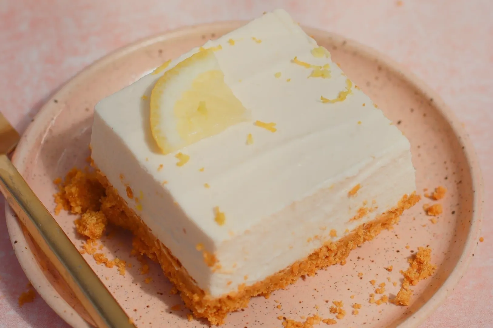 A slice of creamy dessert on a pink plate, garnished with a lemon slice and set on a textured surface.
