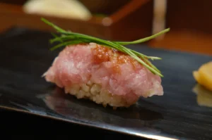 Close-up of sushi with pink tuna tartare on rice, garnished with green chives. Presented on a dark plate, creating a fresh, elegant feel.