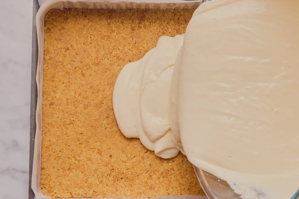 Creamy batter is being poured onto a crumbly graham cracker crust in a square baking pan, set on a marble countertop, suggesting cheesecake preparation.
