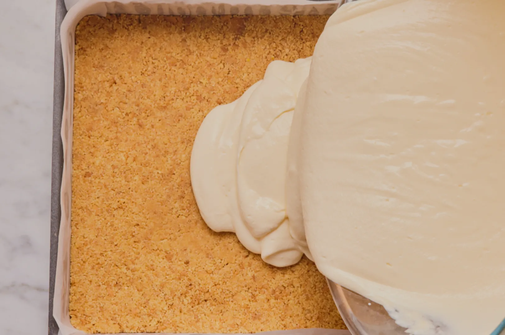 Creamy batter is being poured onto a crumbly graham cracker crust in a square baking pan, set on a marble countertop, suggesting cheesecake preparation.