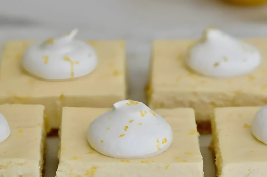 A close-up of cheesecake squares topped with dollops of whipped cream and lemon zest.