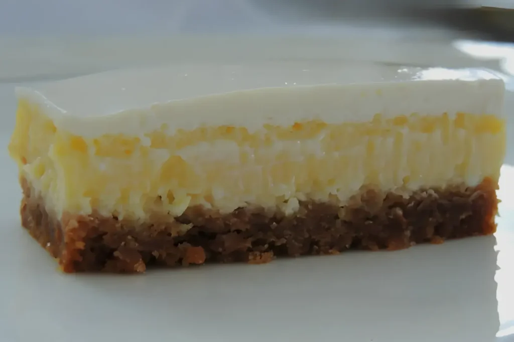 A slice of cheesecake with a graham cracker crust, featuring a creamy layer on top and a slightly visible layer of yellow filling underneath.