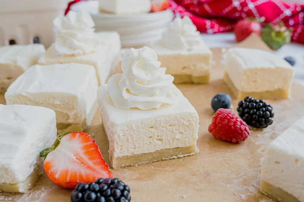 Creamy cheesecake squares topped with whipped cream sit on parchment paper. Fresh strawberries, blackberries, and raspberries add color and freshness.