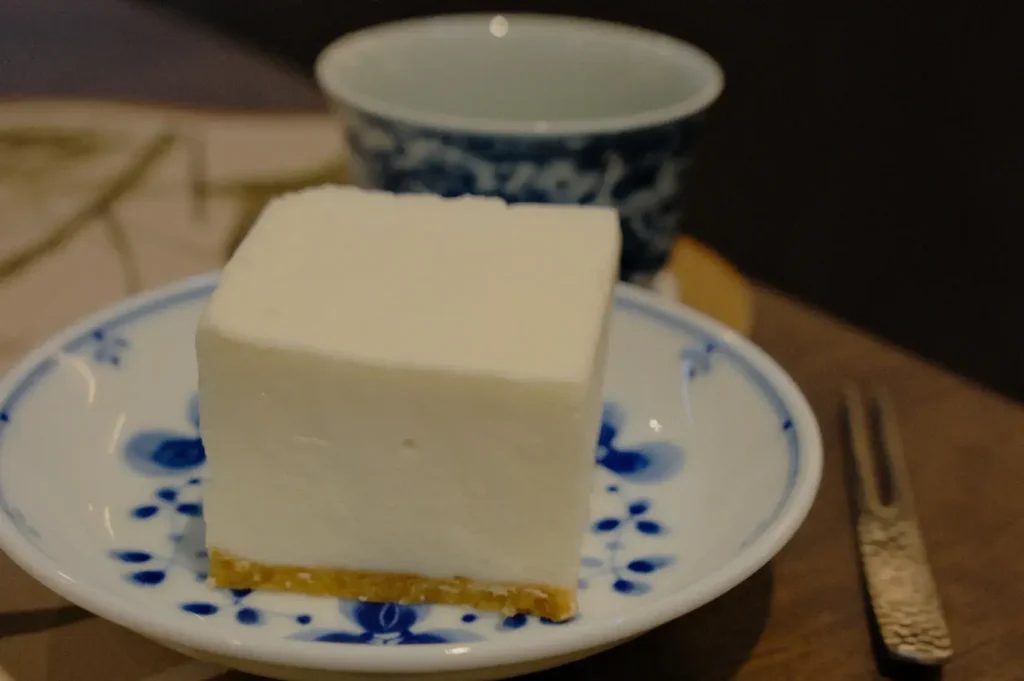 A square piece of white dessert on a blue and white plate, with a cup in the background.