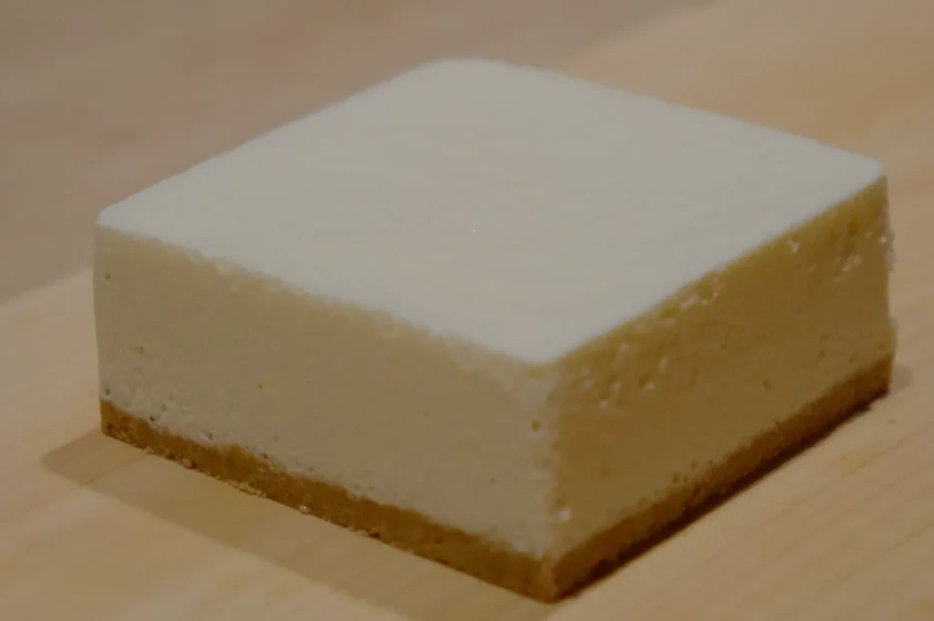 Square slice of creamy cheesecake with a golden brown crust on a wooden surface, conveying a simple and inviting dessert presentation.