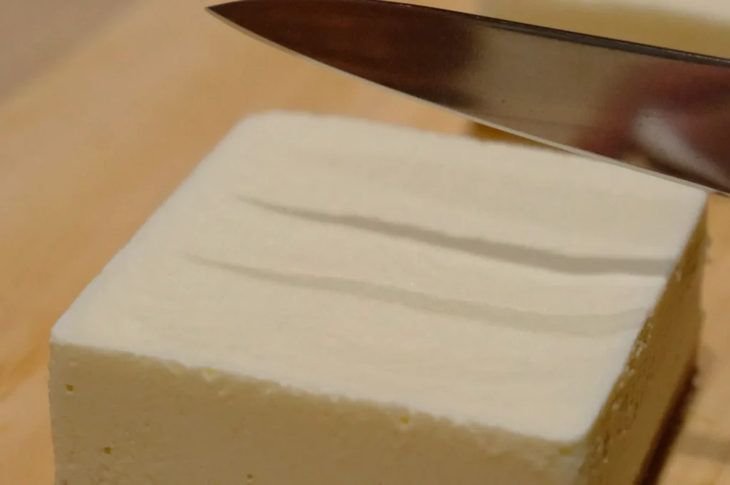 A square soybean cheesecake being sliced with a knife on a wooden cutting board.