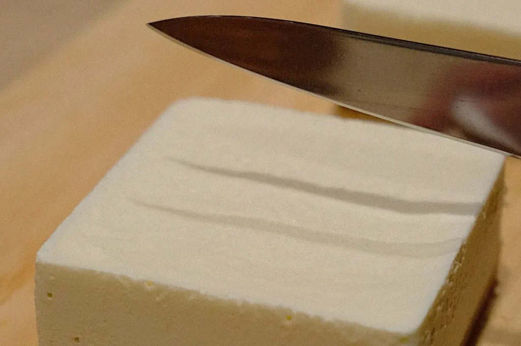 A knife slices through a smooth, white block of tofu on a wooden cutting board. The scene conveys a sense of careful preparation and precision.