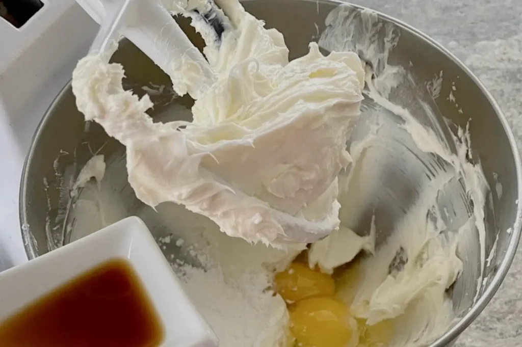 A mixing bowl with whipped cream and egg yolks, alongside a small dish of vanilla extract.