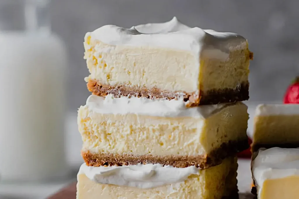 Three layers of cheesecake bars with a creamy top and a graham cracker crust, stacked on a wooden surface, with a glass of milk in the background.