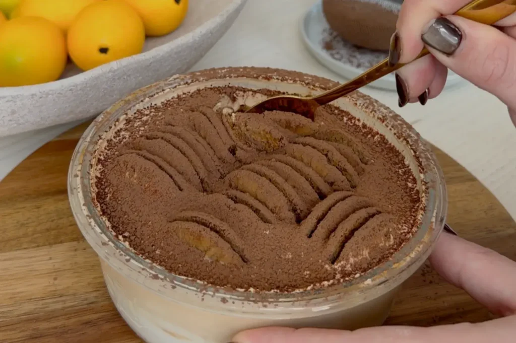 A hand holding a spoon is about to dig into a tiramisu topped with cocoa powder in a glass dish. In the background, there's a bowl of citrus fruits.