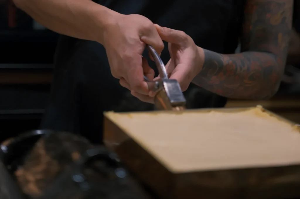 Chef Masa uses a metal tool to shave a rectangular block of cheese. The scene is intimate, focused, with a culinary setting.