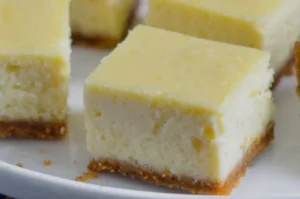Close-up of creamy cheesecake squares with a thick, crumbly graham cracker crust. The soft, moist texture suggests a rich, indulgent dessert.