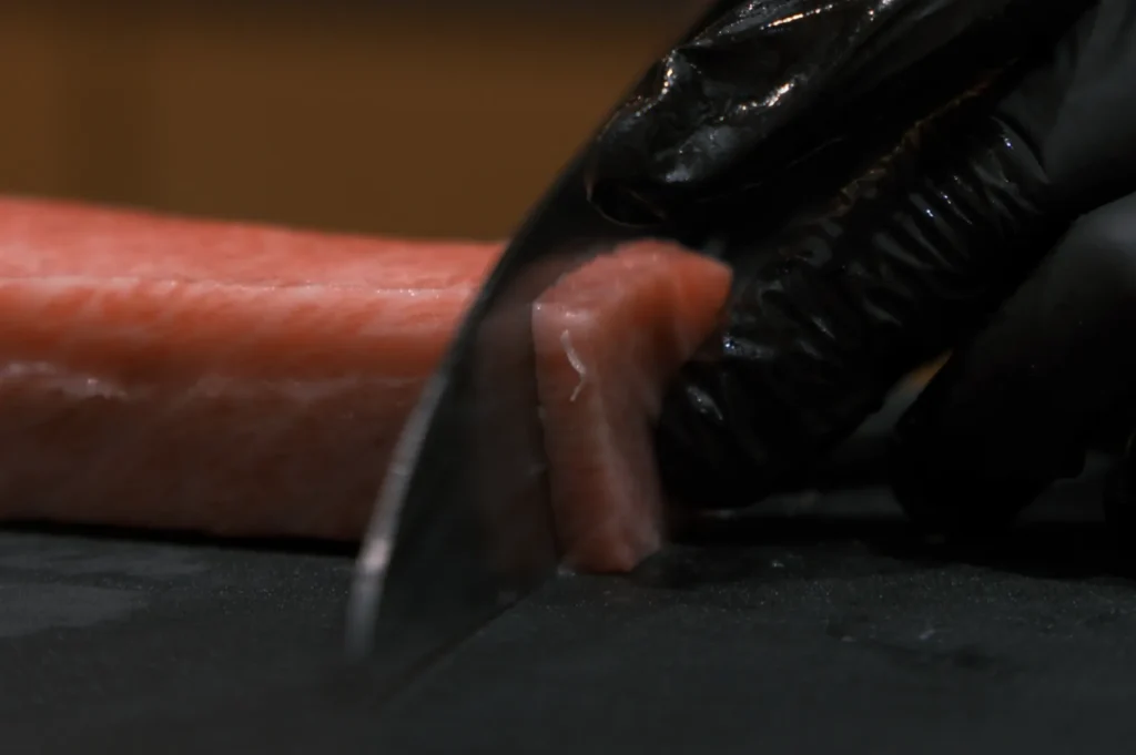 Chef Masa‘s gloved hand slices a piece of raw salmon with a sharp knife on a dark cutting board. The focus is on precision and freshness in a culinary setting.