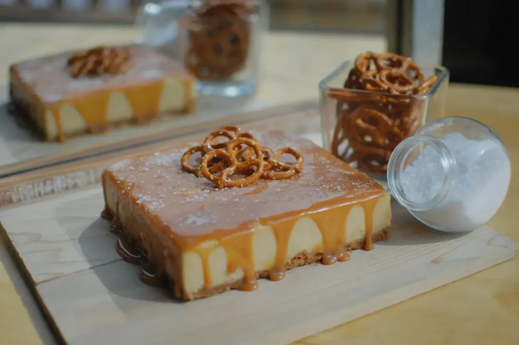 A square caramel dessert topped with pretzels, sitting on a wooden surface, with a small jar of pretzels and a container of salt nearby.