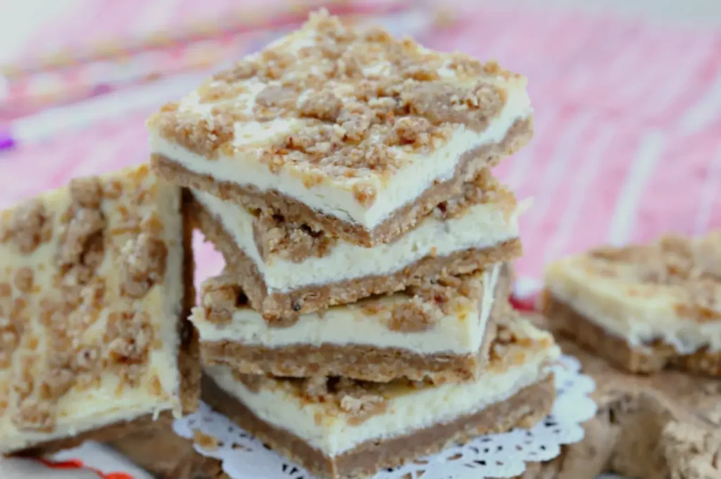 A stack of dessert bars with a creamy layer topped with crumbled toppings, resting on a lace doily on a pink and white striped surface.