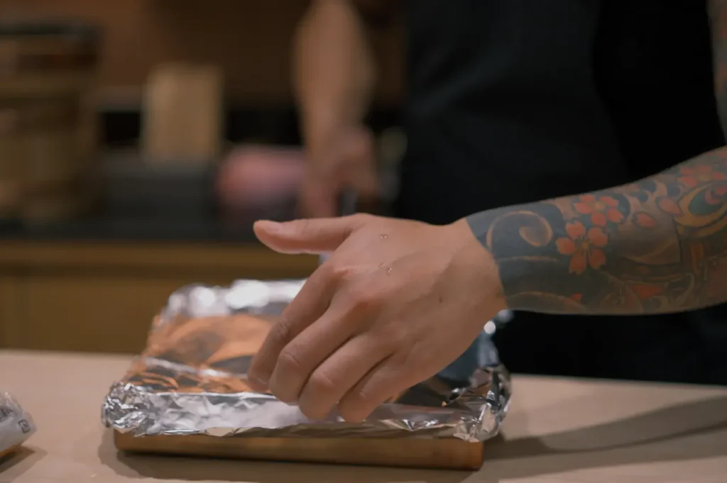 A close-up of Chef Masa's hand placing a foil-covered dish on a wooden surface, with a blurred figure in the background.