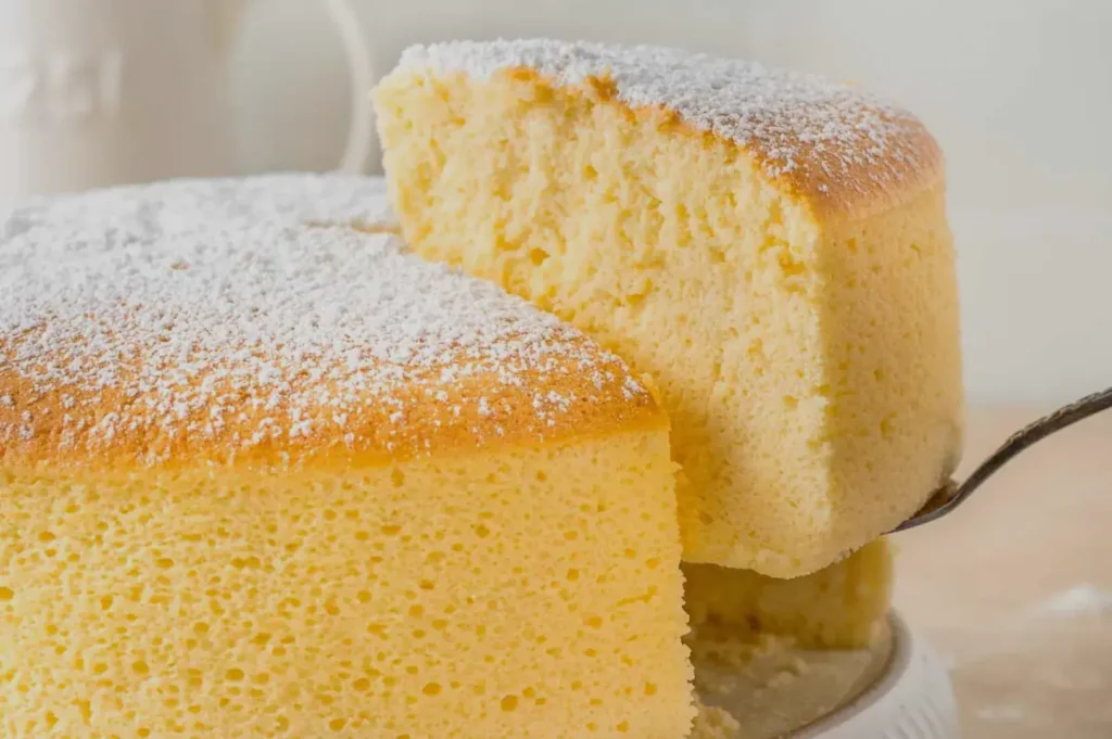 A fluffy, golden-brown Japanese cheesecake topped with powdered sugar is shown, with a slice being lifted on a metal spatula, conveying a light and airy texture.