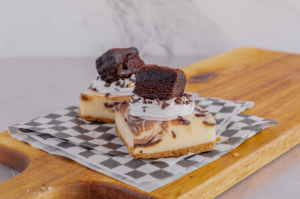 Two marble cheesecake squares topped with whipped cream, chocolate shavings, and a brownie piece sit on a checkered paper on a wooden board.