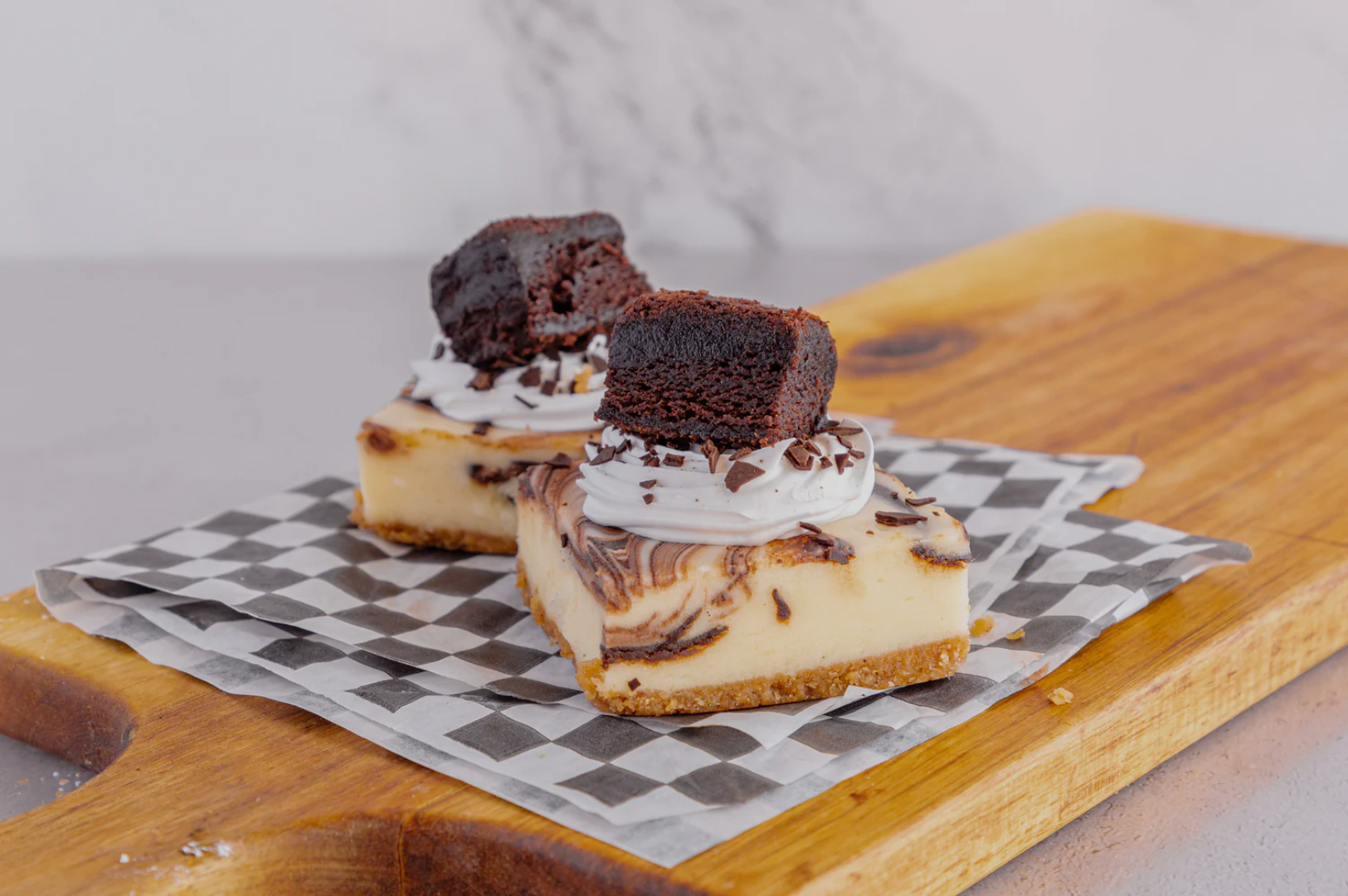 Two marble cheesecake squares topped with whipped cream, chocolate shavings, and a brownie piece sit on a checkered paper on a wooden board.