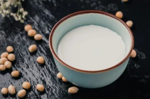 A small blue bowl filled with soy milk, surrounded by scattered yellow soybeans on a dark surface.
