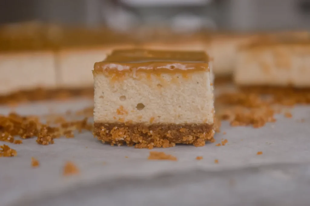 A close-up of a square slice of cheesecake with a smooth caramel topping and a crumbly graham cracker crust, surrounded by crumbs on a white surface.