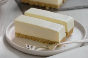 Two rectangular slices of cheesecake on a white plate with a fork beside them. The cheesecake has a smooth, creamy surface and a golden crust.