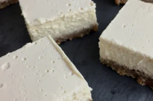 Squares of cheesecake with a graham cracker crust on a dark surface.