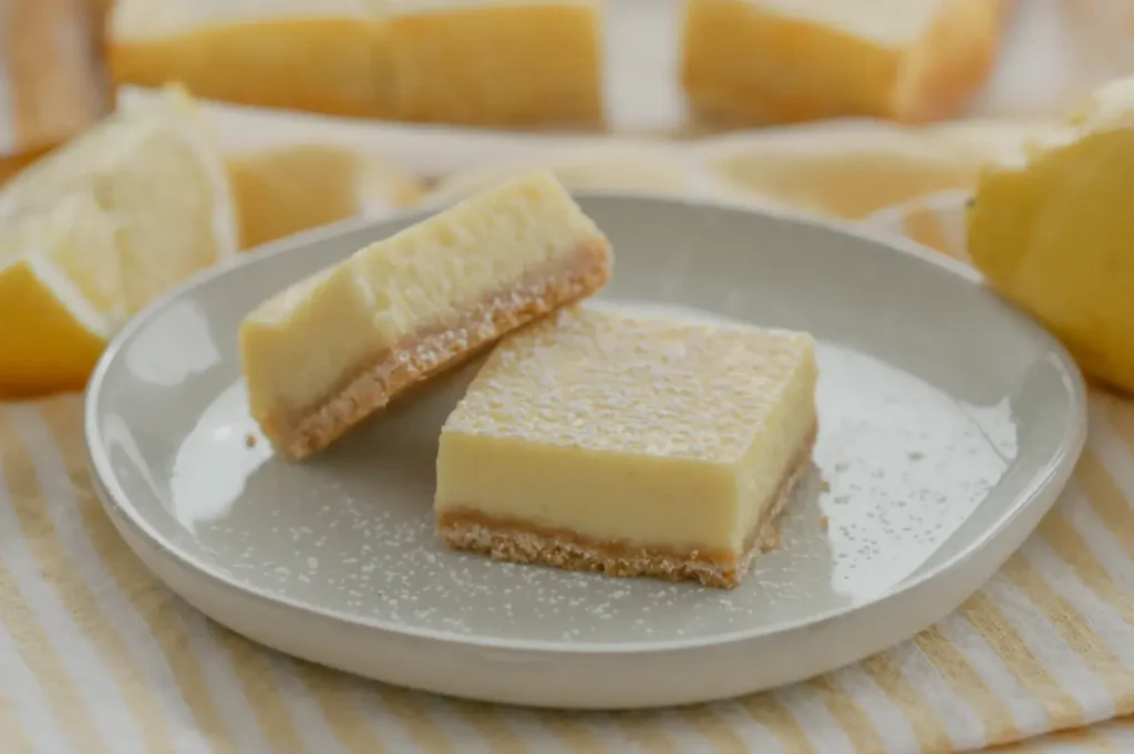 A plate with two lemon bars, one slightly tilted, with a light dusting of powdered sugar, surrounded by lemon slices in the background.