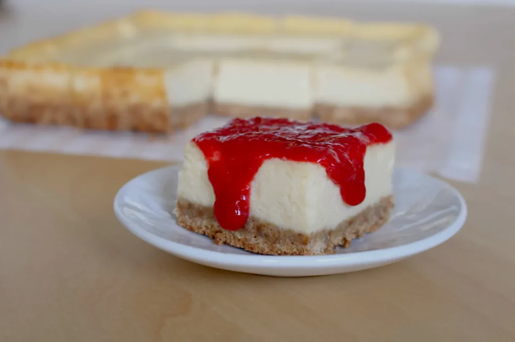 A slice of cheesecake with a red fruit topping on a small white plate, with a larger cheesecake in the background.