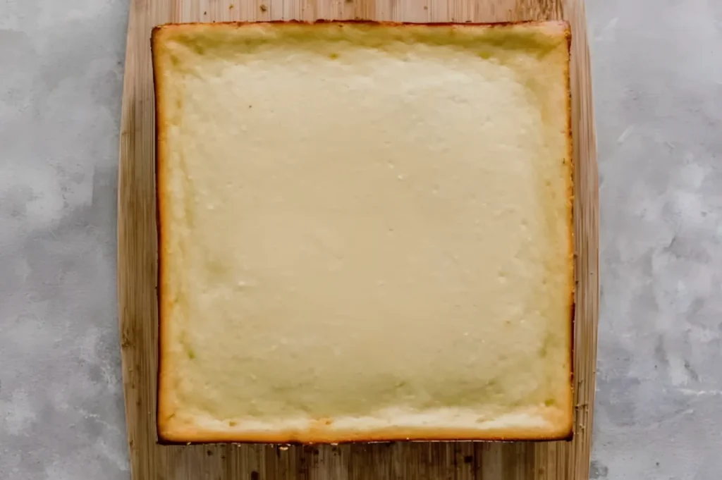 A square cheesecake with a golden-brown edge on a wooden cutting board.