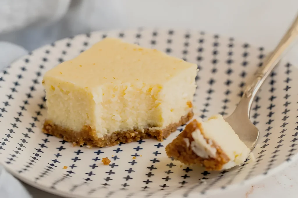 A slice of cheesecake on a patterned plate, with a fork beside it and a small piece cut out.