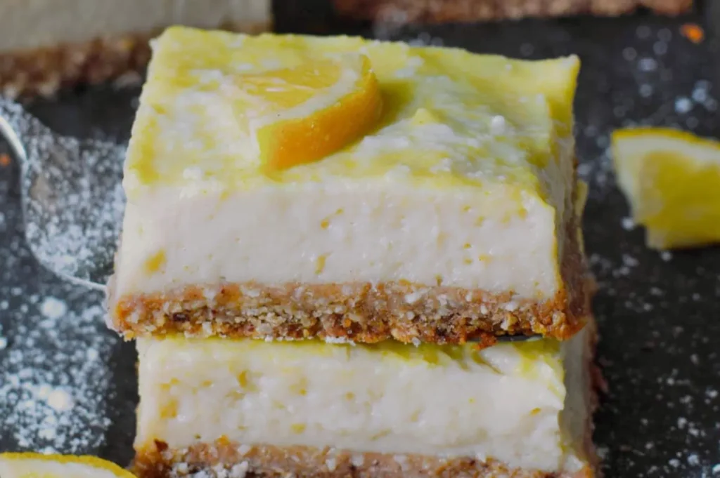 Two layered lemon bars stacked on top of each other, with a glossy lemon glaze and a slice of lemon on top. Dusting of powdered sugar is visible around the bars.