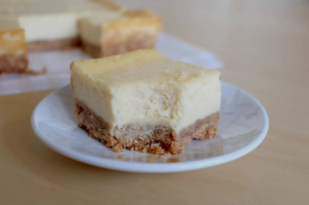 A slice of cheesecake on a small white plate, with a graham cracker crust, sitting on a wooden surface.