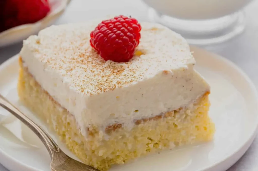 A piece of cake topped with whipped cream and a raspberry on a white plate, with a spoon beside it.