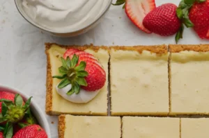 A cheesecake dessert cut into squares, topped with fresh strawberries and a dollop of whipped cream, with additional strawberries in a bowl nearby.