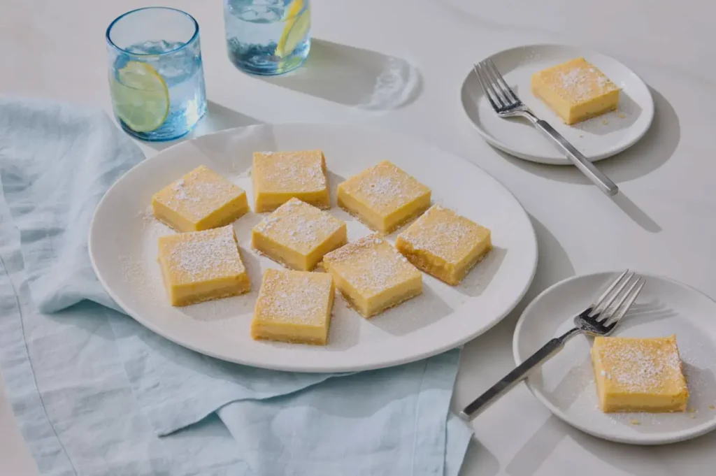 A plate of lemon bars dusted with powdered sugar, surrounded by plates and forks, with glasses of drinks in the background.