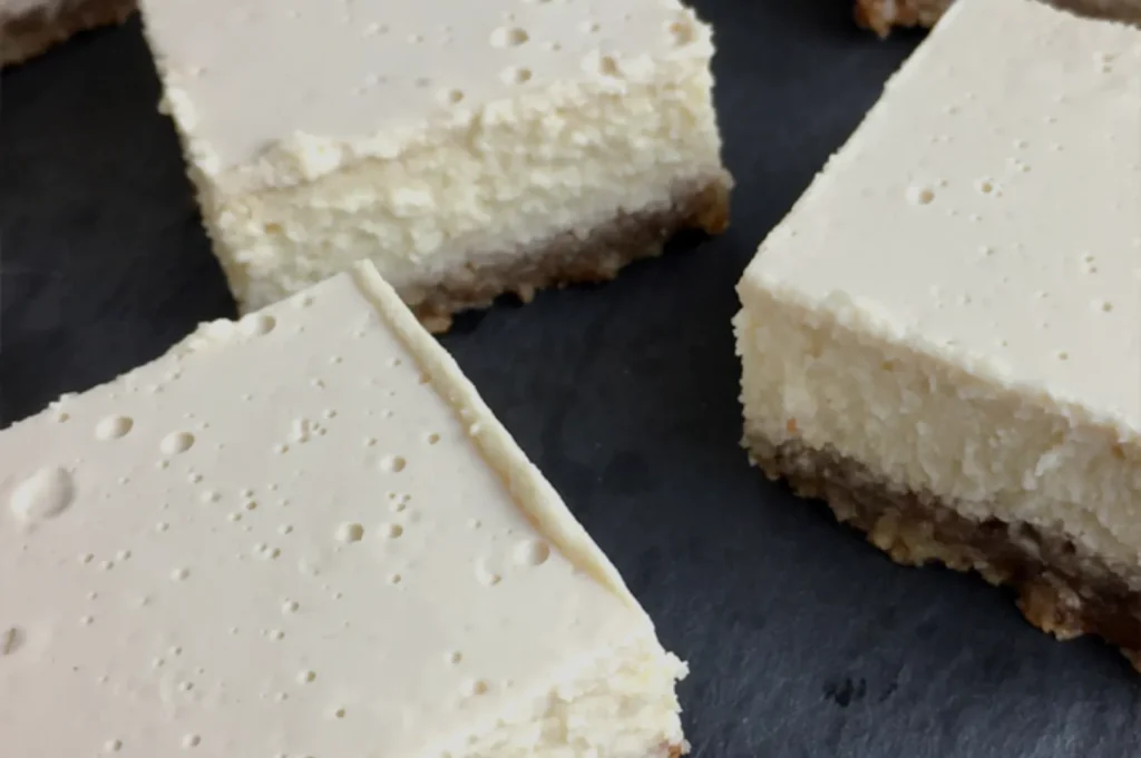 A close-up of cream cheese squares with a crumbly base, arranged on a dark surface.