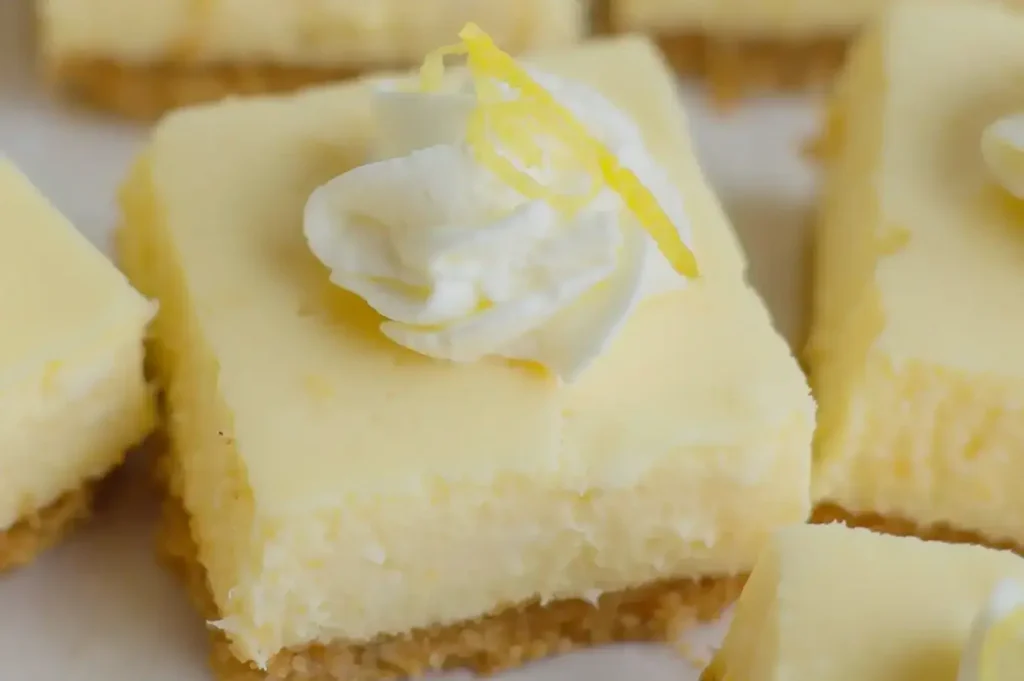 A close-up of lemon cheesecake squares with a swirl of whipped cream and a decorative lemon twist on top.