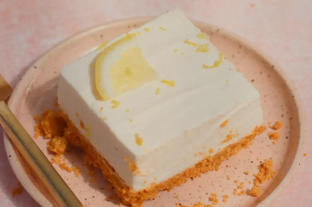 A square dessert with a creamy white topping and a lemon slice on top, on a light pink plate with a golden fork beside it.