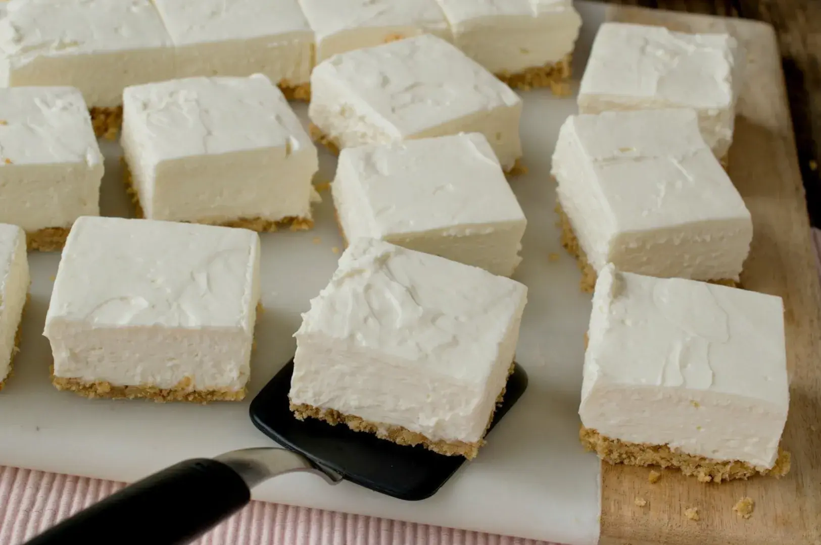 A close-up of square pieces of cheesecake on a cutting board, some pieces are being lifted with a spatula.