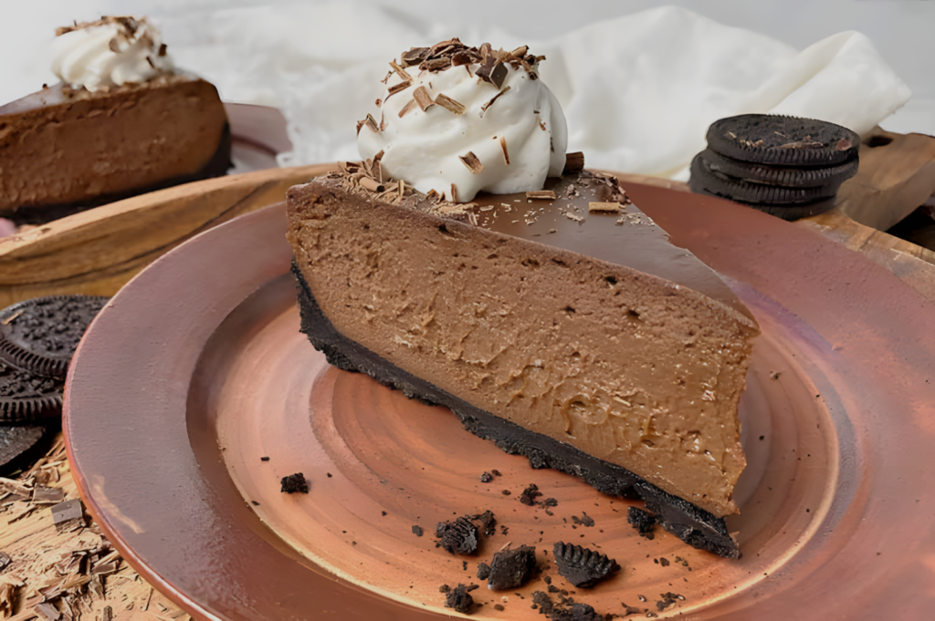 A rich chocolate cheesecake slice on a brown plate with a dark cookie crust, topped with whipped cream and chocolate shavings, set against a cozy backdrop.