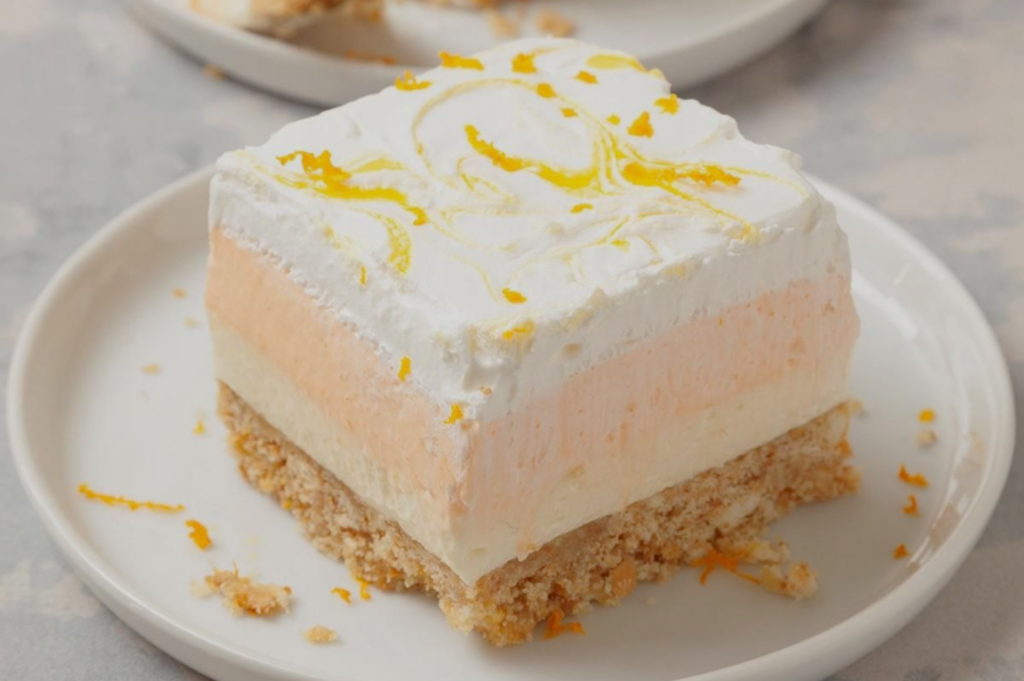 A creamy, layered dessert with a crumbly base on a white plate. It features white and peach layers, topped with whipped cream and orange zest.
