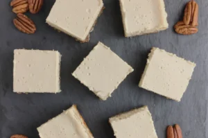 A top-down view of square pieces of dessert with a creamy topping, arranged on a slate surface with scattered pecans around them.