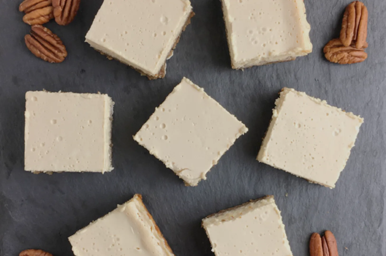 A top-down view of square pieces of dessert with a creamy topping, arranged on a slate surface with scattered pecans around them.