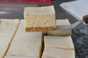 Close-up of a fork lifting a square slice of creamy layered cheesecake, with a crumb crust and more neatly cut pieces on a table.