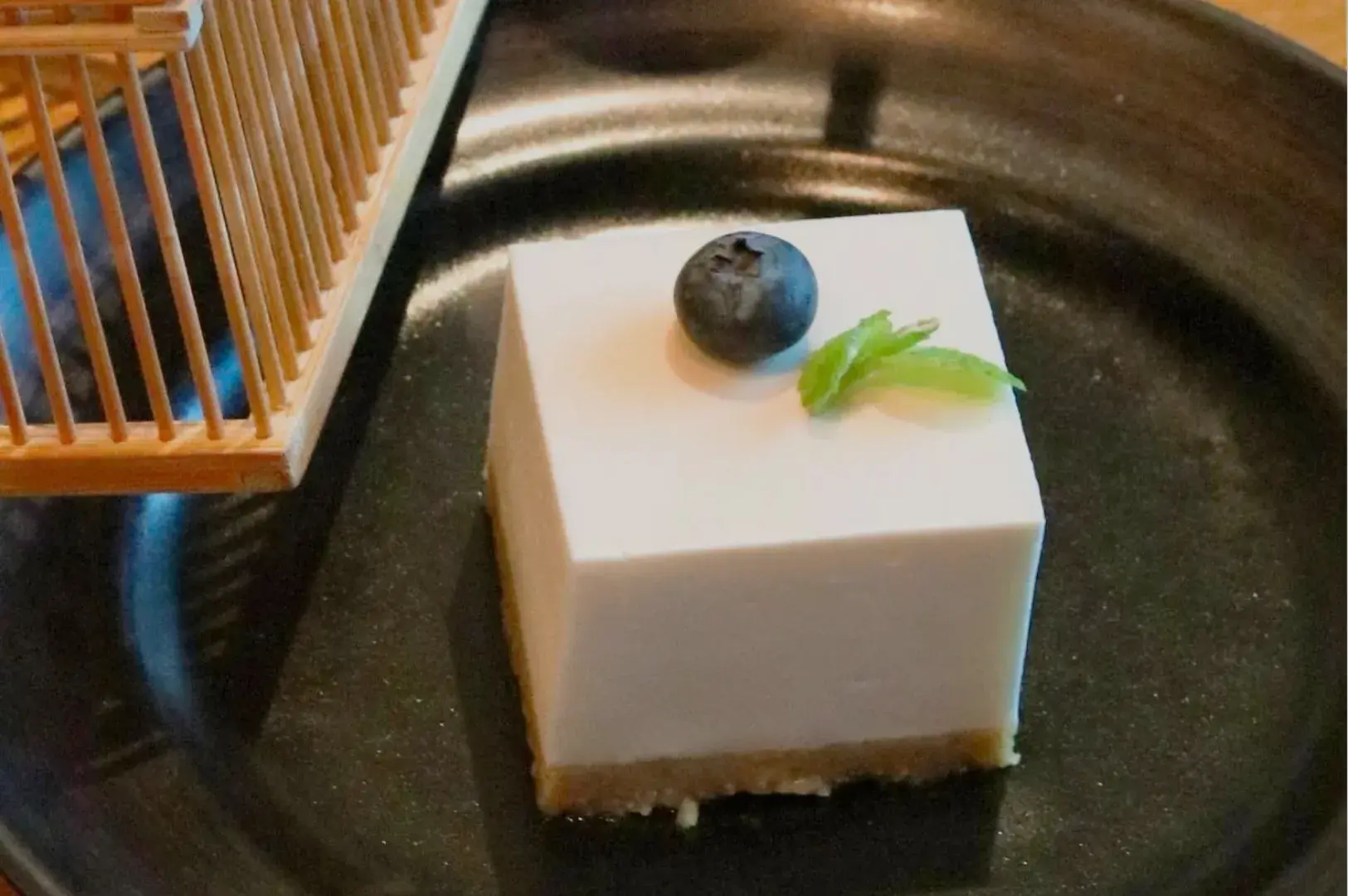 A square slice of cheesecake on a black plate, topped with a blueberry and a small green garnish, with a wooden structure in the background.