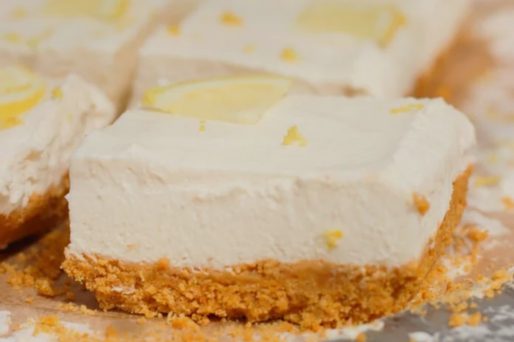 Close-up of creamy lemon bars with a crumbly graham cracker crust. The bars are topped with lemon zest and slices, conveying a fresh, zesty feel.